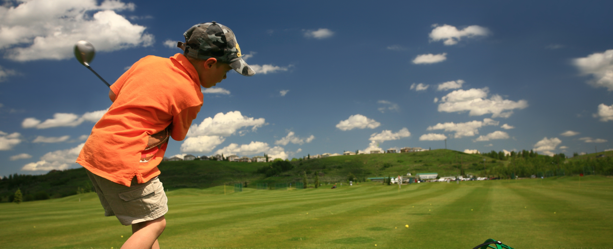 Image of boy in orange polo shirt, swinging a golf club on a golf course